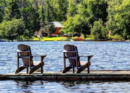 Deck chairs on a dock overlooking a lake with a cabin on the shore.