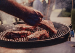 Close-up photo of thick steaks being grilled outside. In the background, friends and family sit in the yard, waiting for a meal.