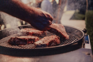 Close-up photo of thick steaks being grilled outside. In the background, friends and family sit in the yard, waiting for a meal.