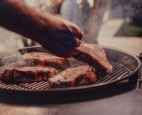 Close-up photo of thick steaks being grilled outside. In the background, friends and family sit in the yard, waiting for a meal.