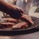 Close-up photo of thick steaks being grilled outside. In the background, friends and family sit in the yard, waiting for a meal.