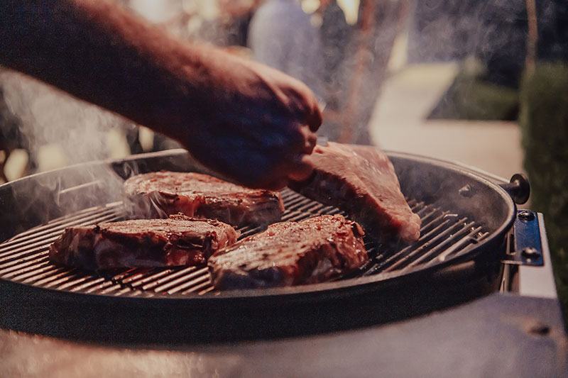 Close-up photo of thick steaks being grilled outside. In the background, friends and family sit in the yard, waiting for a meal.